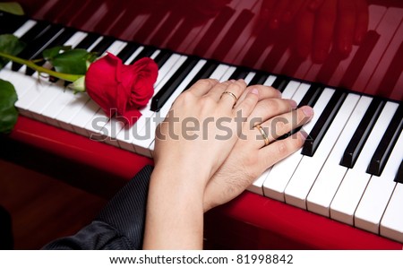 Hands of married man and woman with wedding rings laying on keys of red grand piano with red rose nearby