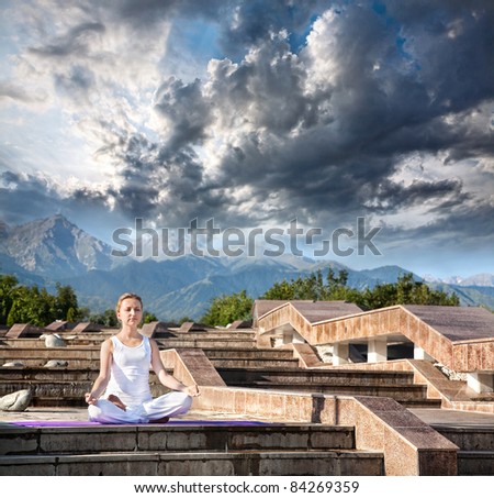Woman doing yoga meditation in white cloth. Ardha padmasana, half lotus pose with dhyana mudra gesture. Woman Sitting on the stone stairs at Mountain and dramatic clouds background