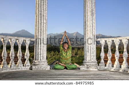 Yoga padmasana lotus pose is done by funny Indian man in green cloth between stone columns at mountain background