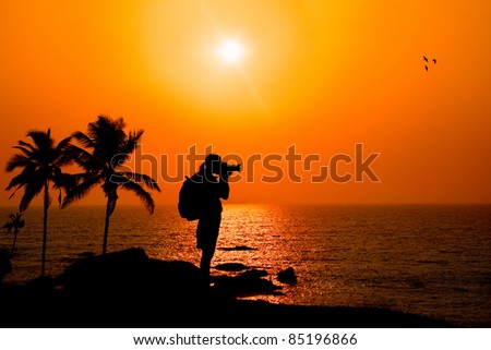 Photographer silhouette shooting sea outdoors on the rock cliff near palm trees at sunset background
