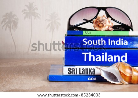 Guide books on the table with Seashells and sunglasses on its at palm trees and ocean background. Books with titles: South India, Bali, Sri Lanka, Goa, Thailand