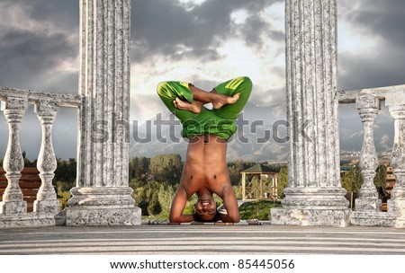 Yoga urdhva padmasana, head standing pose in lotus by smiling Indian man in green trousers near stone column at mountain and dramatic sky background