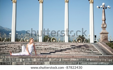 Yoga Vakrasana I twisting pose by Beautiful Caucasian woman in white cloth. Woman sitting on stone stairs and columns, mountains, blue sky at background