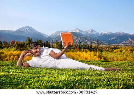 Indian Man in white cloth doing Yoga eka pada shirshasana foot behind the head pose and reading orange book at mountain and blue sky background. Free space for text on the book and sky