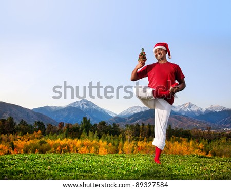 Christmas yoga vrikshasana tree pose by happy Indian man in white trousers, red socks and Christmas hat with Christmas tree at mountain background. Free space for text