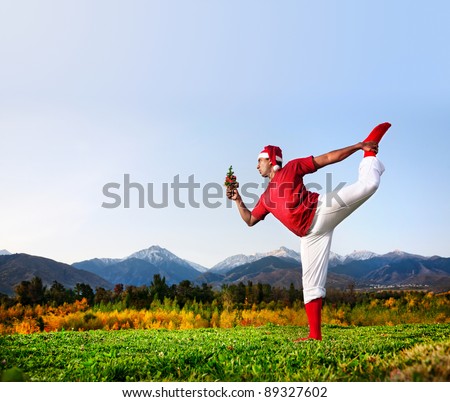 Christmas yoga natarajasana dancer pose by funny Indian man in white trousers, red socks and Christmas hat with looking at Christmas tree at mountain background. Free space for text