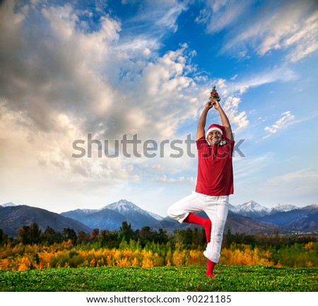 Christmas yoga vrikshasana tree pose by happy Indian man in white trousers, red socks and Christmas hat with Christmas tree at mountain background. Free space for text