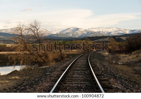 Railroad tracks vanishing around a curve with snow covered mountains, a pond, and green trees in the middle