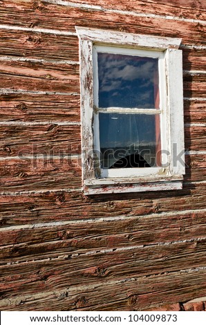 Window with a broken pane in an old, weathered,  log building shows reflections of clouds and mountains with snow in the remaining glass