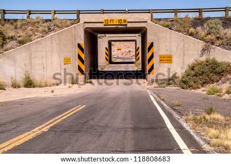One lane tunnel under a highway bridge with a sharp curve at the end blocking the view of oncoming traffic.  There are plenty of warning and caution signs.