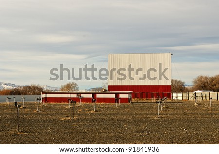 Drive-in theater with a bright white and red screen, dirt parking area, and speakers on posts just waiting for warmer weather