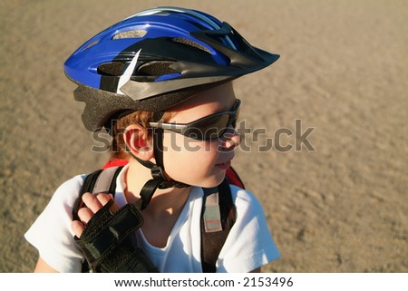 A nine year old boy wearing sunglasses, a bike helmet and a backpack crouches in a sunny park.