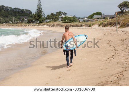 A male surfer seen from behind walking on the beach of Matapouri, New Zealand, with his board in one hand