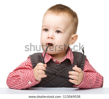Cute little child play with book and biting glasses while sitting at table, isolated over white