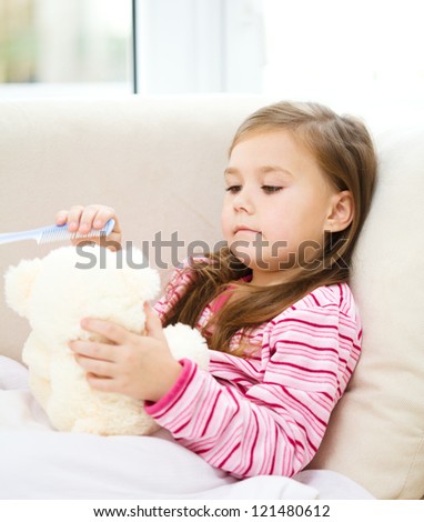 Little girl is brushing her teddy bear while laying in bed and wearing pajama, indoor shoot