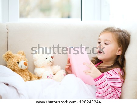 Little girl is reading a story for her teddy bears while laying in bed and wearing pajama, indoor shoot