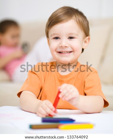 Little boy is drawing on white paper using crayon, his sister sits behind him
