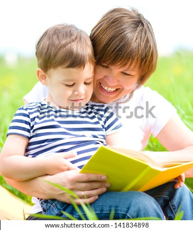 Mother is reading book for her child while sitting on a green grass outdoors