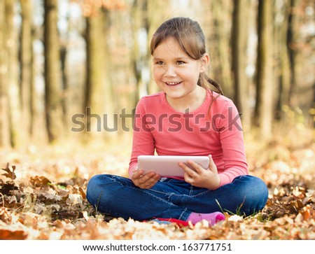 Little girl is reading from tablet while sitting on yellow autumn leaves, outdoor shoot