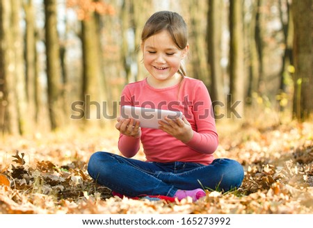 Little girl is reading from tablet while sitting on yellow autumn leaves, outdoor shoot