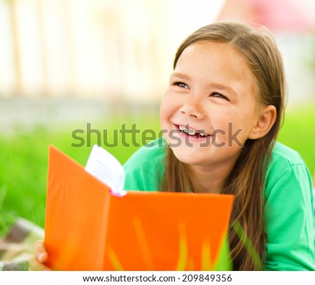 Cute little girl is reading a book while laying on green grass