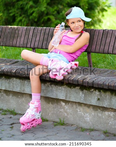 Little girl is wearing roller-blades in city park and drinking mineral water