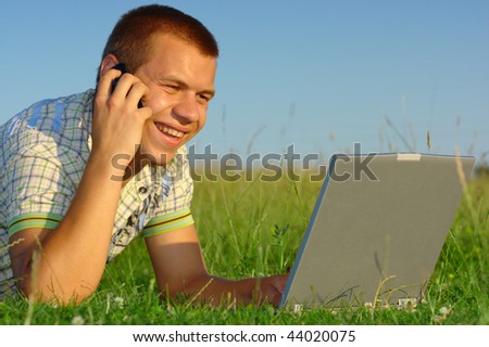 Happy student working with laptop and talking to a cell phone on green meadow, summer sunny day