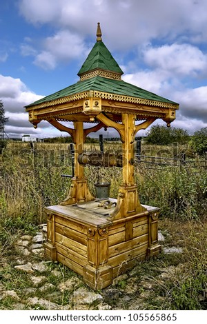 Wood draw-well with a green roof and doors, a bucket on a background of grass, bushes, sky
