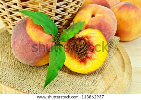 Several whole peaches, half peach, twig with green leaves, wicker basket on a napkin of burlap and wooden round board