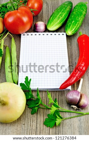 Small notepad, red tomatoes, pod red pepper, parsley, garlic, onion, two cucumbers, green bean pods on an old wooden board