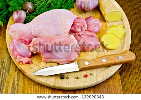 Cut pieces of chicken legs on a round board, garlic, parsley, ginger, dill, pepper and a knife on a wooden boards background