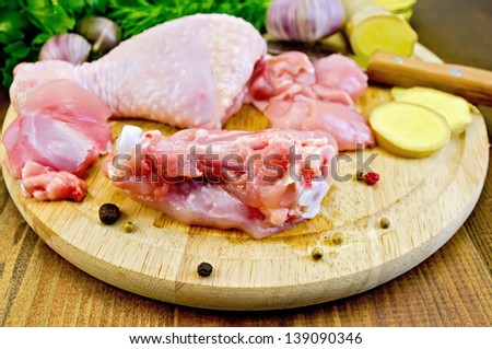 Chicken leg cut on a round board, garlic, parsley, ginger, dill, pepper and a knife on a wooden boards background