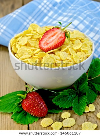 Cornflakes in a white bowl with half a strawberry, leaves and berries of a strawberry, a napkin on a wooden boards background