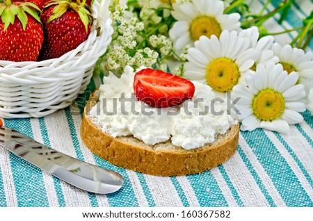 Hunk of bread with cottage cheese cream and strawberries, a basket with berries, a knife, a bouquet of daisies on a green striped cloth