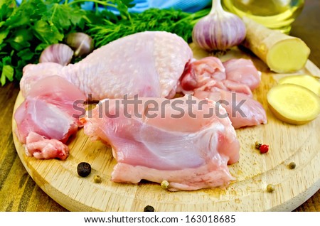 Cut pieces of chicken legs on a round board, garlic, parsley, ginger, dill, oil and knife on a wooden board