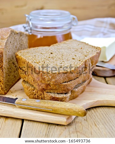 A stack of slices of rye homemade bread with a knife on a plate, napkin, loaf of bread, a jar of honey on a wooden board