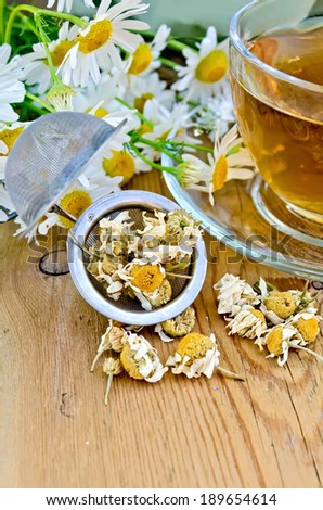 Herbal tea in a glass cup, metal sieve with dry chamomile flowers, fresh flowers, daisies, doily on a wooden board