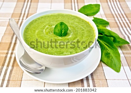 Cream soup green from spinach in a white bowl and saucer, spoon, spinach leaves on the background fabric