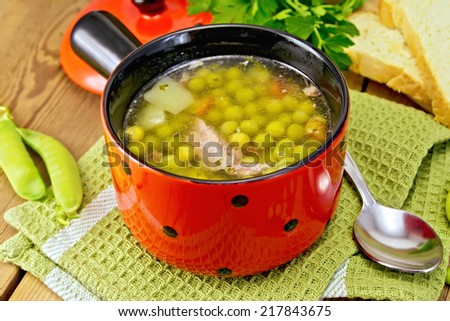 Green pea soup with meat and potatoes in a red bowl, bread, spoon, parsley on a green napkin on the background of wooden boards