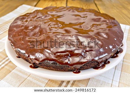 Round chocolate cake with chocolate icing on a plate on a napkin on a wooden boards background