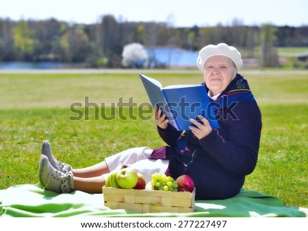 Senior happy woman sitting on a blanket on glade in the park. She is reading book. Healthy outdoor activities. Happy and smiling.