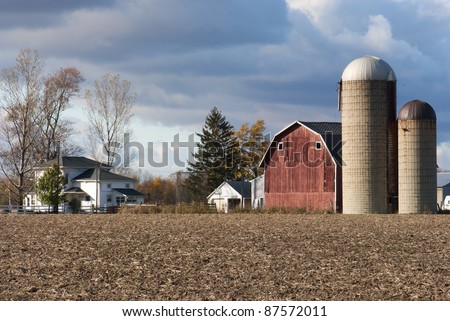 Farm yard with old barn and two silos. A plowed field in the foreground and cloudy sky above.
