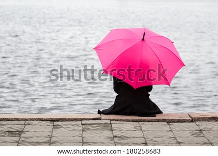Woman sitting lonely with pink umbrella. Concept of taking a break, solitude and thinking with good copyspace on water.