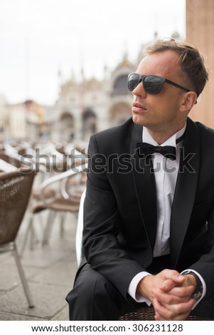 Vertical photo of young handsome man in tuxedo sitting in outdoor cafe