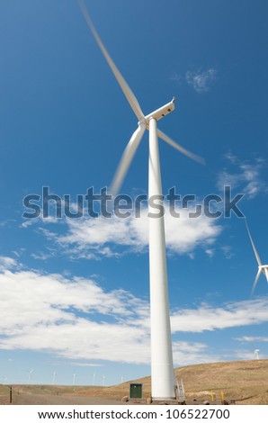 A giant white windmill in Eastern Washington with fast moving blades blurred in motion against a blue sky and some light clouds.