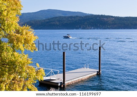 A small boat on a blue lake during early fall with changing trees by a floating dock and mountains in the distance.