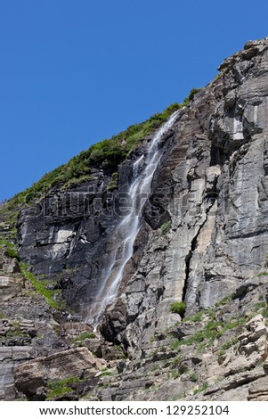 A waterfall created by snow melt cascades over a rock cliff face on a sunny day in Glacier National Park, Montana.