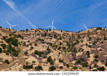 Looking up a steep hillside sparsely covered in trees to giant white windmills on the ridge with a blue sky background.