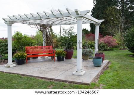 A large white wooden gazebo structure on a concrete platform and a red bench underneath in a landscaped yard.