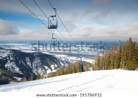 A ski lift going up a steep mountain covered with fresh snow and a valley behind with morning fog.